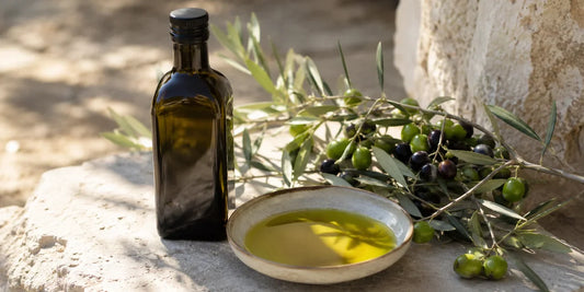 Dark glass olive oil bottle beside fresh green olives and a dish of rich golden-green oil on a pale stone surface