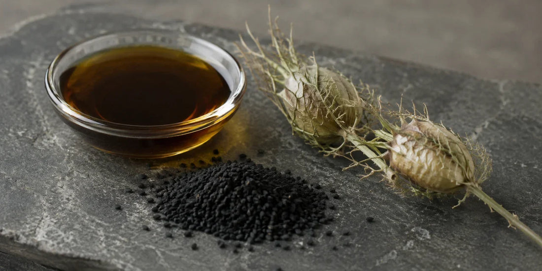A dish of deep bronze black seed oil beside black cumin seeds and dried nigella pods on slate