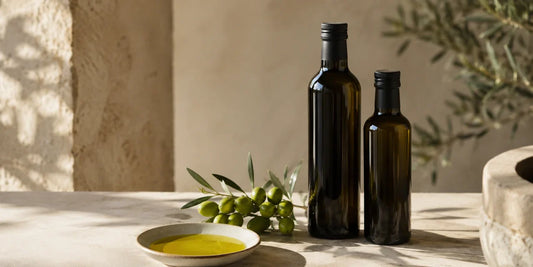 Two dark glass olive oil bottles beside fresh green olives and a dish of rich golden-green oil on pale stone in warm light