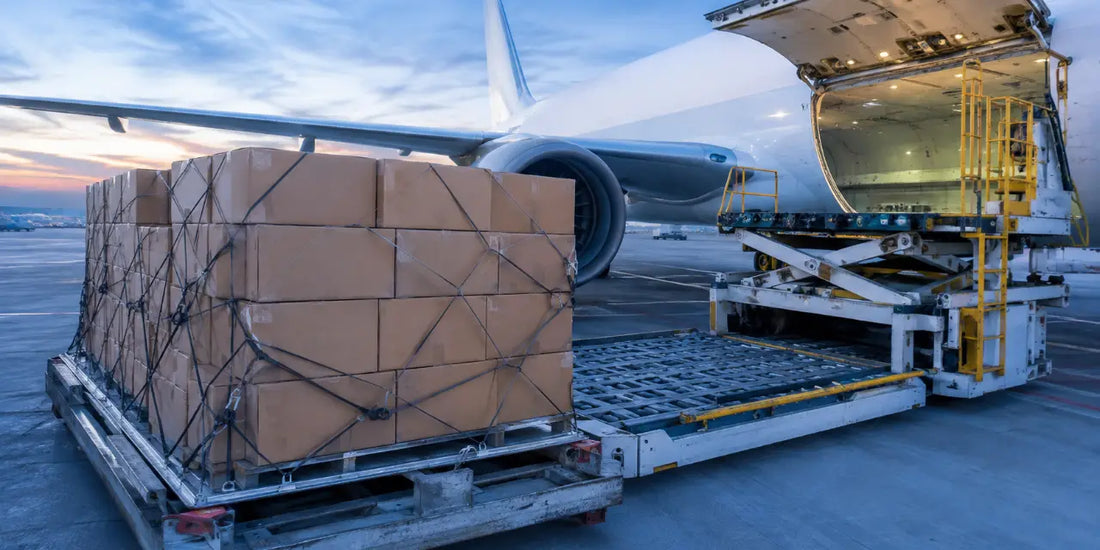 Stacked cardboard cartons of bottles on a pallet beside an aircraft cargo hold at dawn