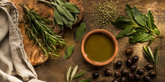 Fresh rosemary, sage and bay leaves beside dried oregano and a dish of olive oil on wood