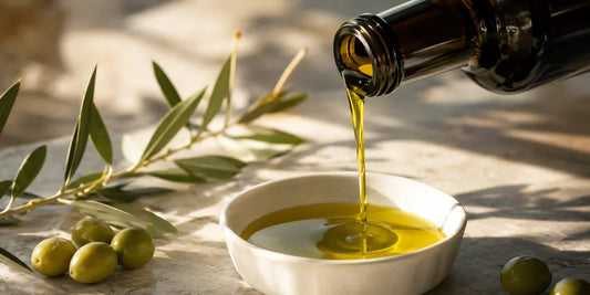Rich golden-green olive oil being poured from a dark glass bottle into a small dish on a pale stone surface in warm daylight