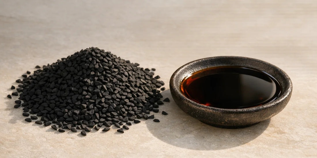 Heap of matte black Nigella sativa seeds beside a dark dish of deep bronze oil on pale stone