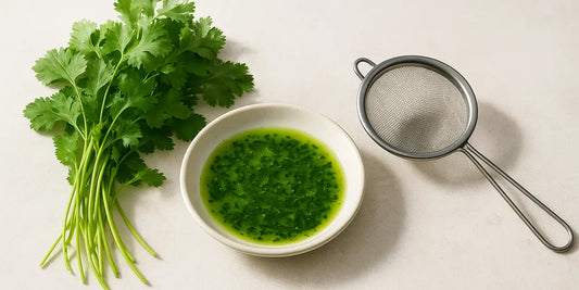 Loose bunch of fresh coriander leaves beside a shallow dish of vivid green herb oil and a fine mesh strainer on pale stone