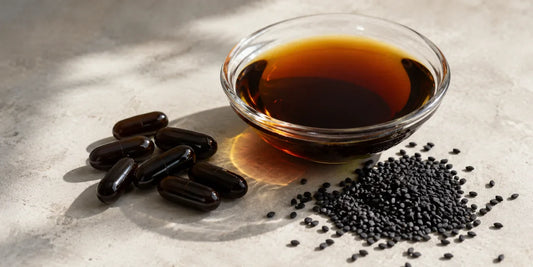 Dark supplement capsules beside a glass dish of deep amber black seed oil and matte black seeds on pale stone