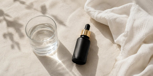 Small clear glass of water beside a matte black bottle of oil and a folded cotton muslin cloth on a pale linen surface in soft daylight