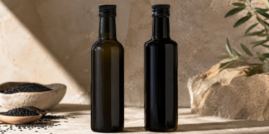 Two dark glass black seed oil bottles side by side on a pale stone surface in warm directional light