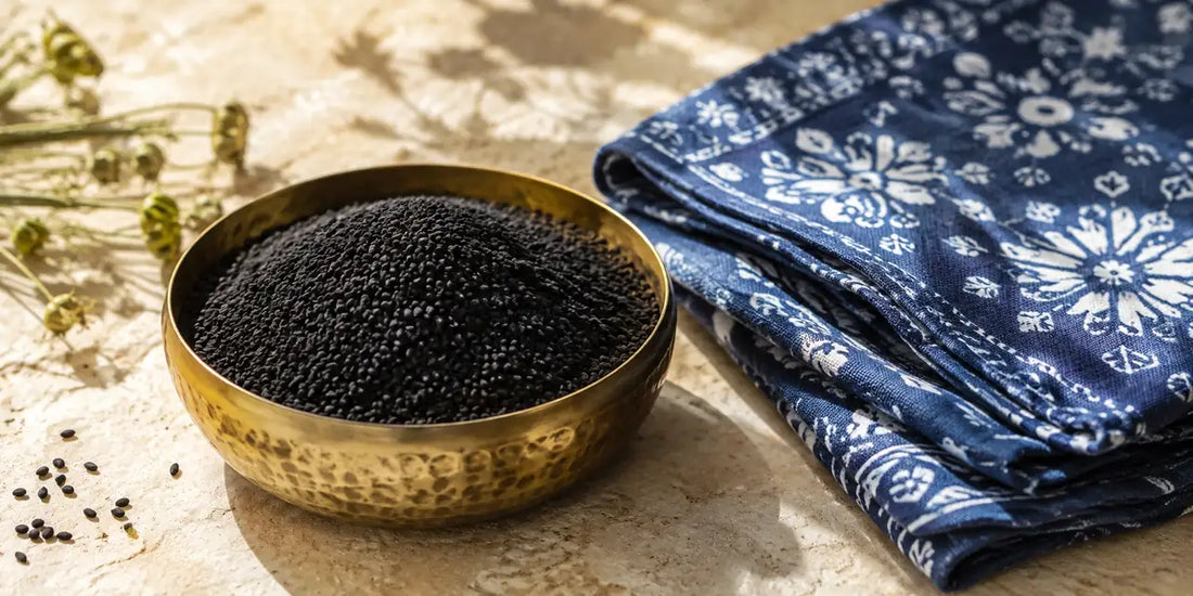 Hammered brass bowl heaped with matte black kalonji seeds beside folded indigo block-printed cloth on warm pale limestone