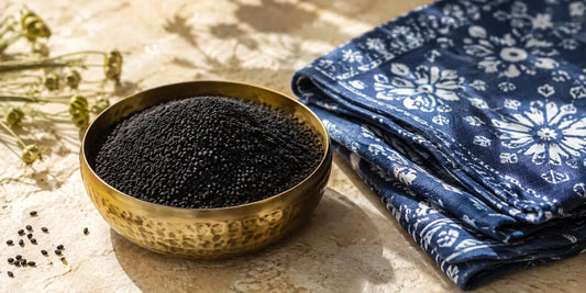 Hammered brass bowl heaped with matte black kalonji seeds beside folded indigo block-printed cloth on warm pale limestone