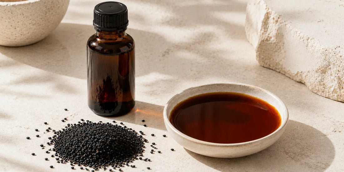 A dark glass black seed oil bottle beside scattered matte black cumin seeds and a small dish of deep bronze oil on a bright pale stone surface