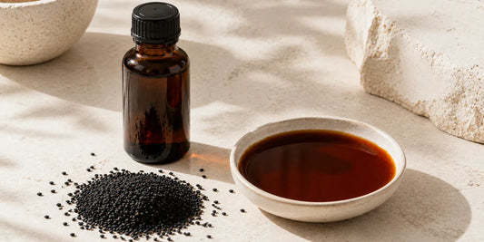 A dark glass black seed oil bottle beside scattered matte black cumin seeds and a small dish of deep bronze oil on a bright pale stone surface