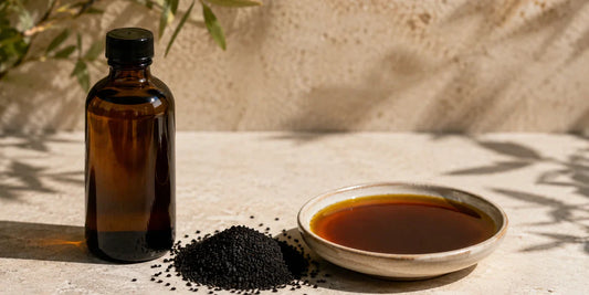 A dark glass black seed oil bottle beside scattered matte black cumin seeds and a small dish of deep bronze oil on a pale stone surface in warm directional daylight