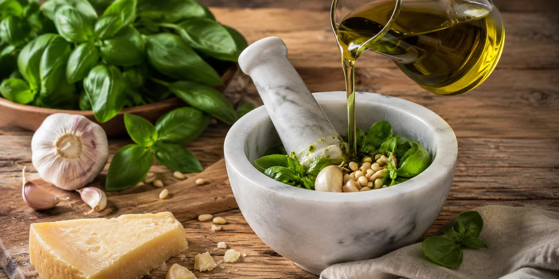 A mortar and pestle of fresh basil pesto with golden-green olive oil pouring in, pine nuts and garlic