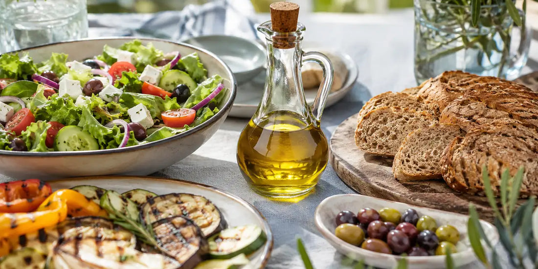 A Mediterranean lunch table with salad, grilled vegetables, bread, olives, and a cruet of olive oil at the centre