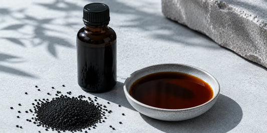 A dark glass black seed oil bottle beside scattered matte black cumin seeds and a small dish of deep bronze oil on a cool pale stone surface