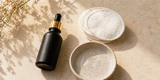 Matte black glass bottle of oil beside soft white cotton pads and a ceramic dish of water on a pale surface