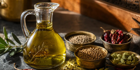 Glass cruet of rich golden-green olive oil beside brass bowls of whole Indian spices on dark slate