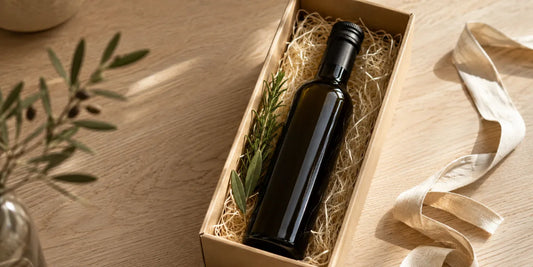 Open kraft gift box lined with wood wool holding a dark glass olive oil bottle and rosemary sprig