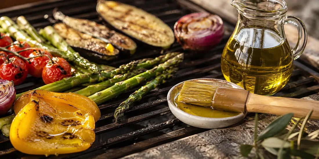 Grilled vegetables beside a jug of golden-green olive oil and a basting brush on a clean grill grate in warm light