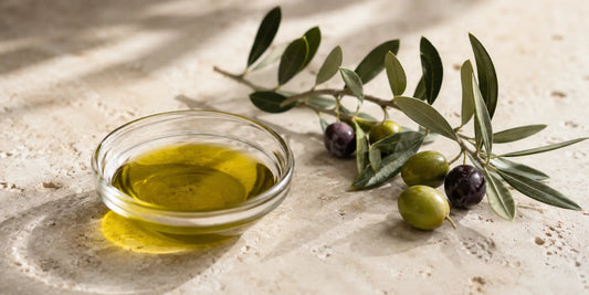 A small glass dish of golden-green olive oil beside a fresh olive branch on a pale stone surface in soft natural light