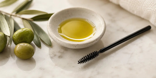 A clean spoolie brush beside a small dish of golden-green olive oil on a marble vanity with olives