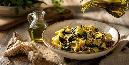 A generous pour of golden-green olive oil over a hearty bowl of pasta with bread on a rustic wooden table in warm light