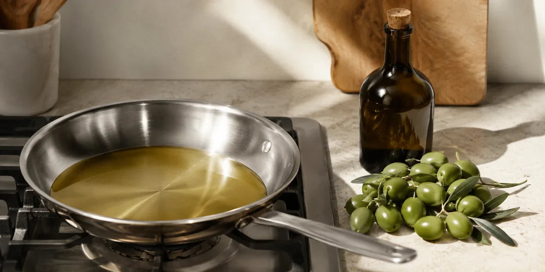 A pan of pale refined olive oil heating on a hob beside fresh olives and a dark glass bottle on stone