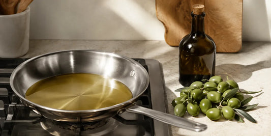 A pan of pale refined olive oil heating on a hob beside fresh olives and a dark glass bottle on stone