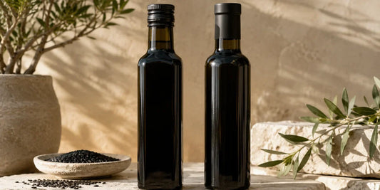 Two dark glass black seed oil bottles side by side on a pale stone surface in warm directional light