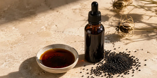Dark glass bottle of deep bronze black seed oil beside scattered matte black seeds on pale stone