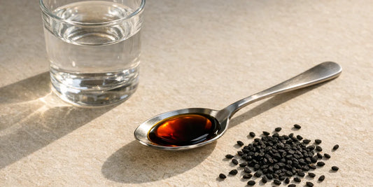 Teaspoon of deep bronze black seed oil beside a small glass of water and scattered black seeds