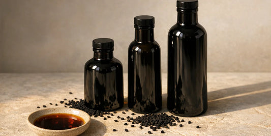 Three graduated dark glass bottles beside scattered black seeds and a dish of deep bronze oil on stone