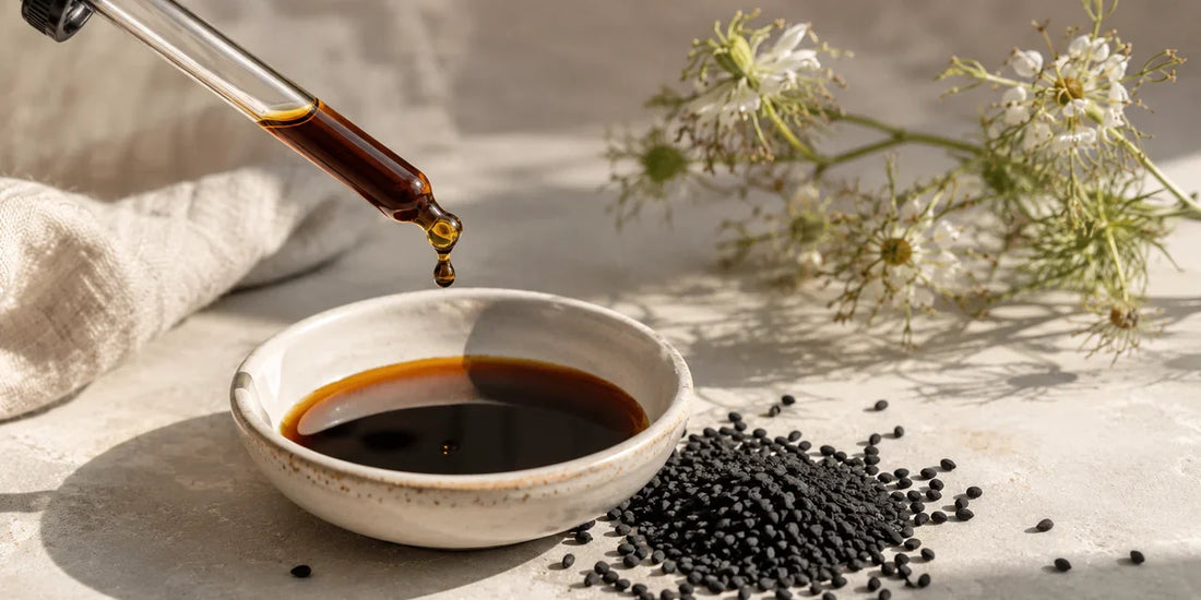 Glass dropper releasing a deep amber black seed oil drop above a small dish, beside matte black seeds on pale stone