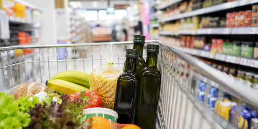 A shopping trolley in a supermarket aisle holding dark green olive oil bottles among groceries