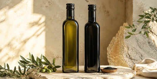 A dark glass bottle of olive oil and a matte black bottle of black seed oil side by side on a pale stone surface in warm directional light