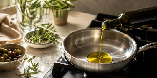 Rich golden-green extra virgin olive oil pouring from a dark glass bottle into a stainless steel pan beside fresh herbs and olives