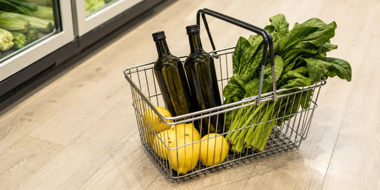Wire shopping basket on a pale wooden floor holding dark glass olive oil bottles, lemons and leafy greens
