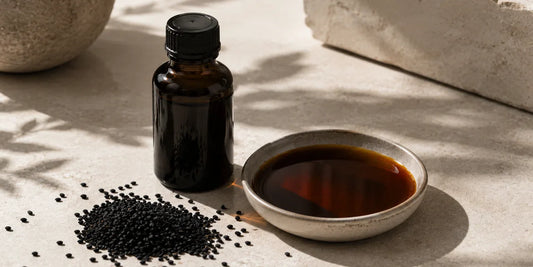 A dark glass black seed oil bottle beside scattered matte black cumin seeds and a small dish of deep bronze oil on a soft pale stone surface