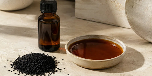 A dark glass black seed oil bottle beside scattered matte black cumin seeds and a small dish of deep bronze oil on a warm stone surface